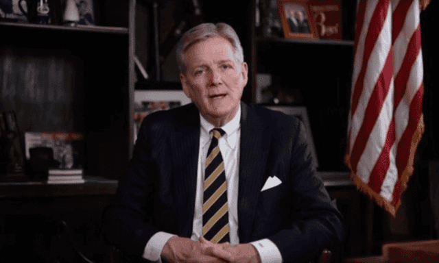 Edwin Brant Frost IV seated at a desk in a dark suit and striped tie, with an American flag behind him