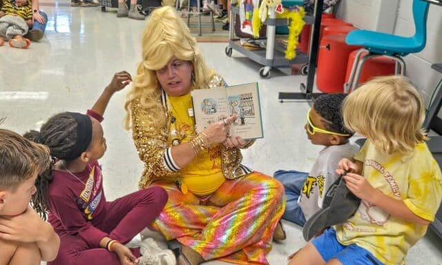 A Crabapple Lane Elementary staff member dressed as Dolly Parton reads a book to students seated on the floor
