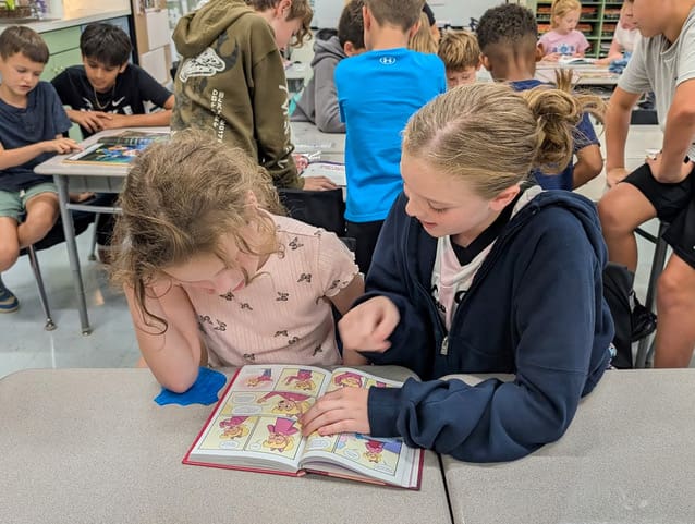 An older student and a younger student read a book together at a table