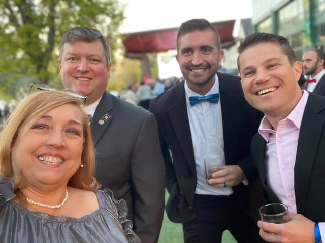 Four guests in formal wear smile for a selfie at a past Bourbon, Bow Ties, Pinot & Pearls event