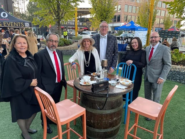 Guests in formal attire gather around a barrel table at a past Bourbon, Bow Ties, Pinot & Pearls event