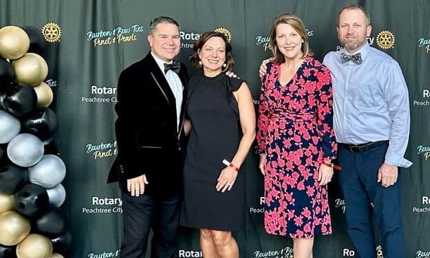 Four guests in formal attire pose in front of the Rotary Club of Peachtree City Bourbon & Bow Ties, Pinot & Pearls backdrop