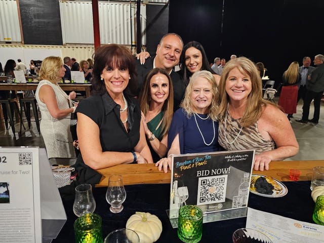 Six guests smile at a table with a silent auction bid sign at a past Bourbon, Bow Ties, Pinot & Pearls event